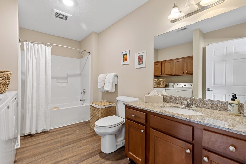 Modern bathroom with wood flooring, granite countertop, single sink, toilet, bathtub with shower curtain, and washer-dryer visible through open door. Neutral-colored walls and simple decor.