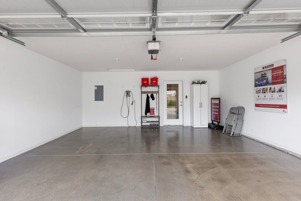 Empty two-car garage with clean, polished floor, wall-mounted storage shelves, cabinets, an electrical panel, some folded chairs, and informational posters on the wall.