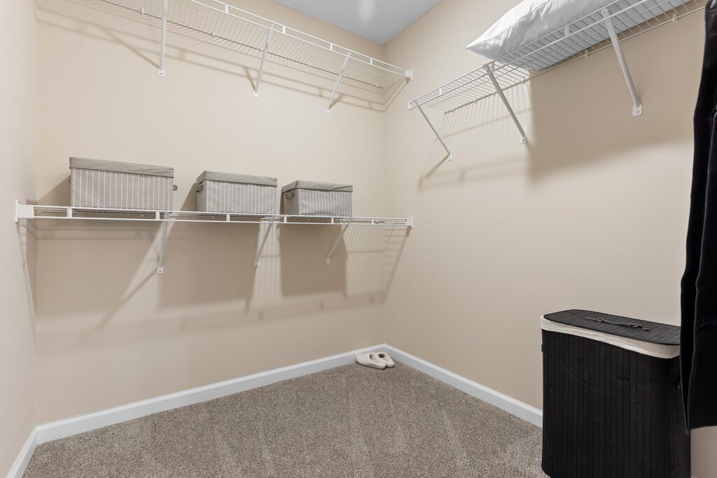 Empty walk-in closet with beige walls, wire shelves holding three storage baskets and a pillow, a black laundry hamper, and carpeted floor with slippers.
