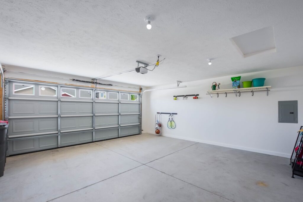 Clean, empty two-car garage with a closed gray door, white walls, ceiling lights, and shelves holding various household items and tools.
