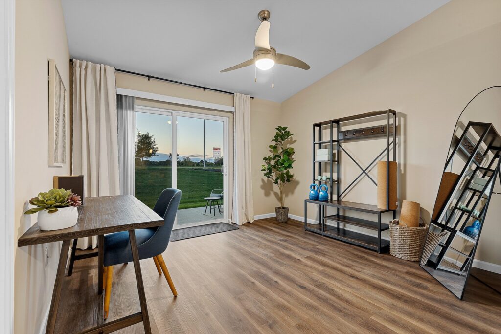 A home office with a wooden desk, blue chair, shelving unit, tall mirror, potted plant, and sliding glass doors opening to a patio with a view of grass and sunset.