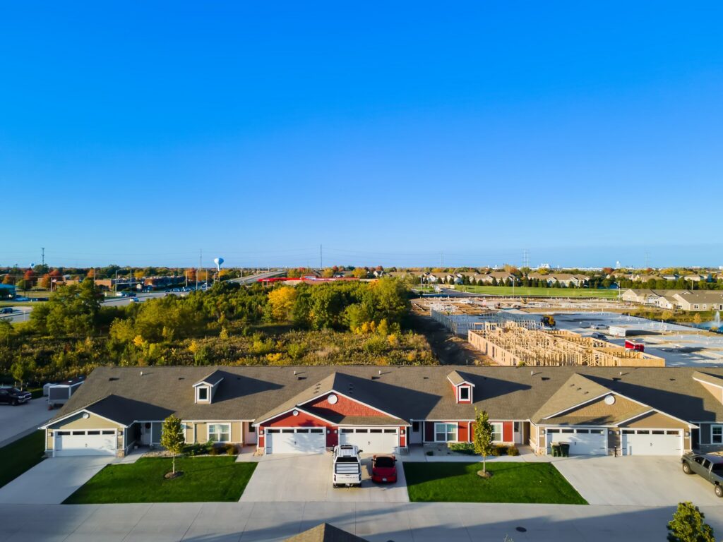 A row of townhouses with garages and driveways faces a construction site and green space under a clear blue sky.