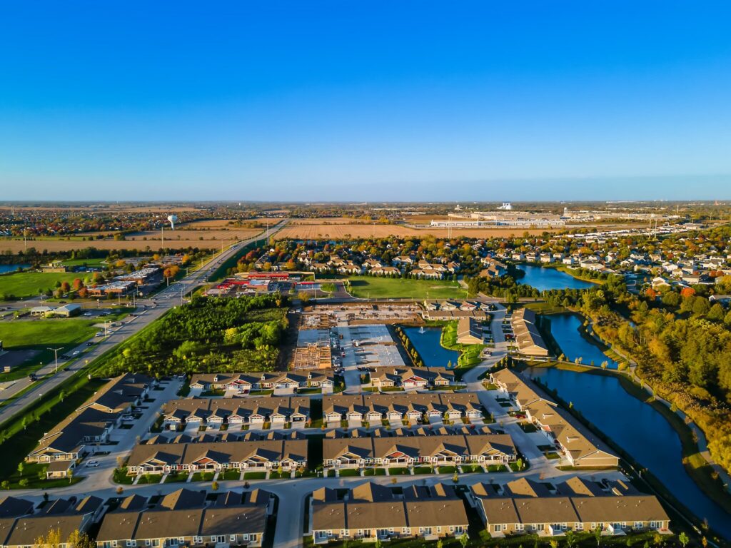 Aerial view of a suburban neighborhood with houses, roads, waterways, green areas, and open land under a clear blue sky.