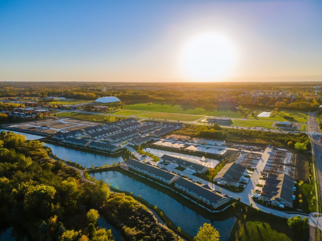 Aerial view of a residential neighborhood with water features and green spaces at sunset, with a bright sun low on the horizon.