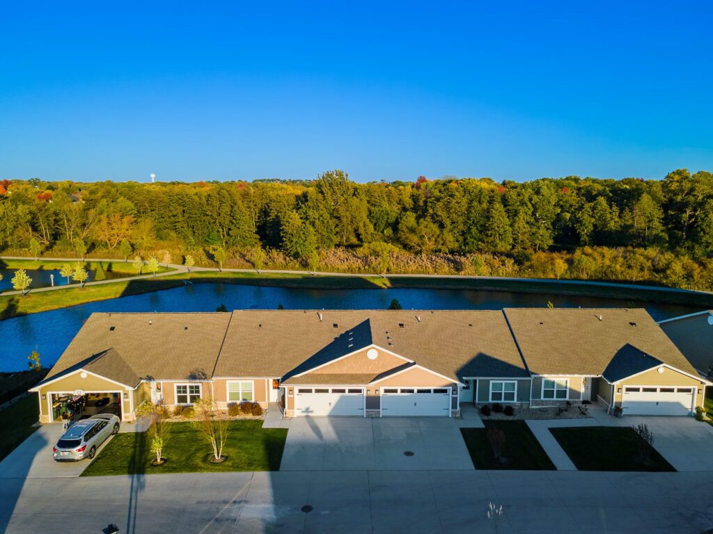 A row of single-story townhomes with attached garages faces a small pond, surrounded by greenery and trees under a clear blue sky.