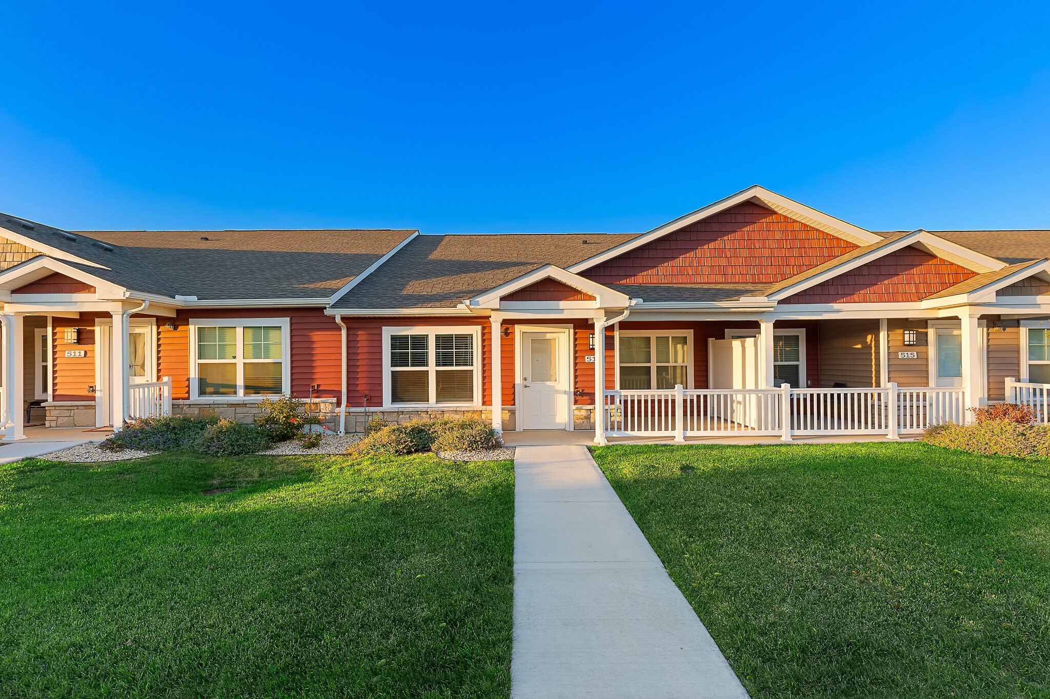 Single-story townhouse with a front porch, red siding, white trim, and a sidewalk leading to the entrance, set against a clear blue sky.