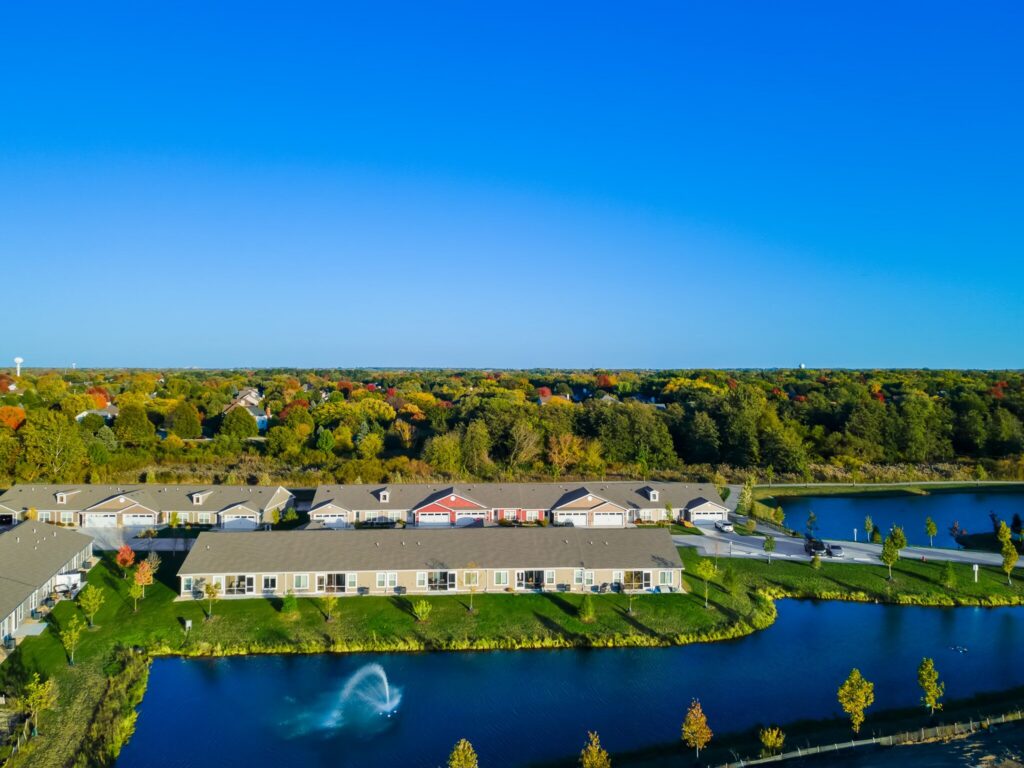 Aerial view of residential buildings near a pond with a water fountain, surrounded by trees under a clear blue sky.