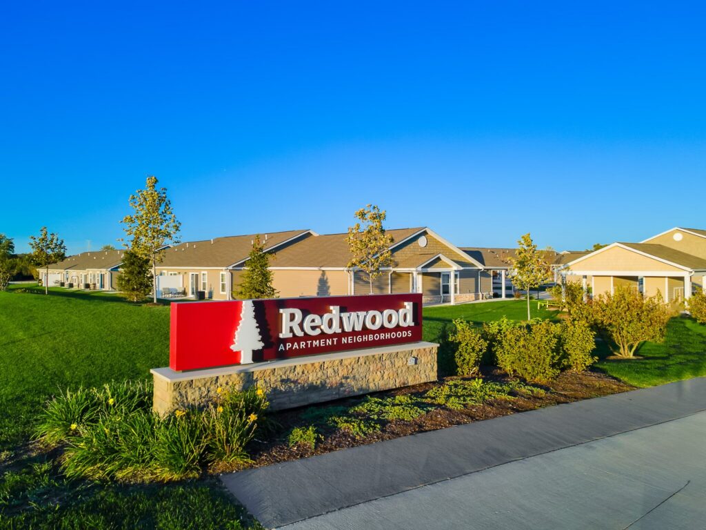 Redwood Apartment Neighborhoods sign in front of single-story residential buildings, with green grass, trees, and a clear blue sky.