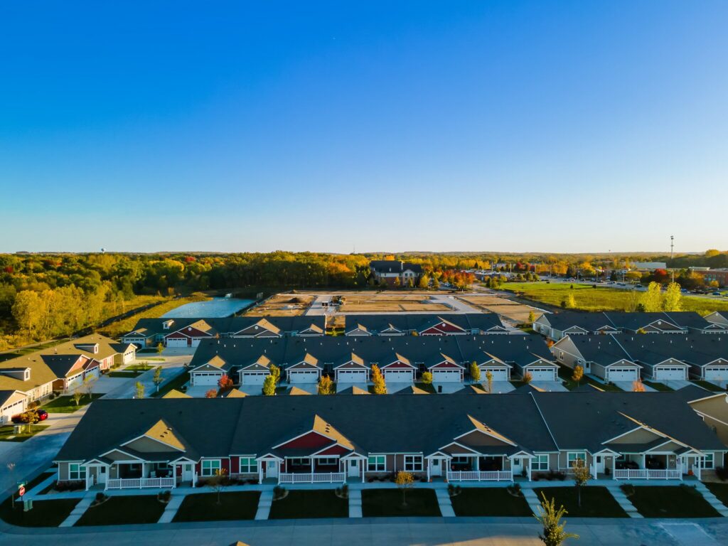Aerial view of a suburban neighborhood with rows of single-story houses, green lawns, trees, and a clear blue sky in the background.