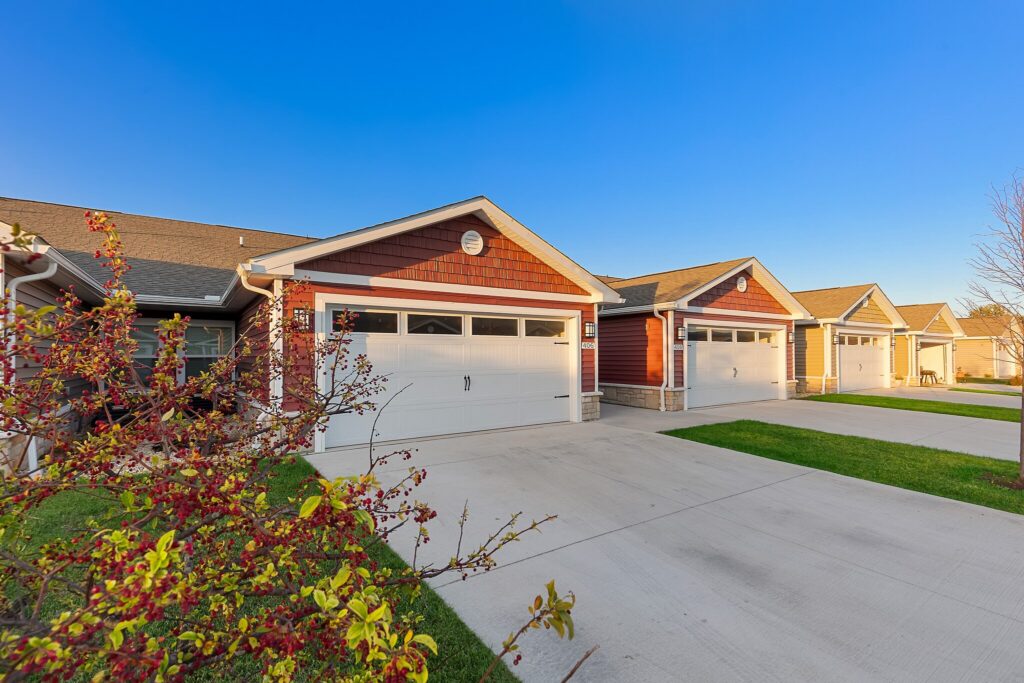 Row of modern suburban homes with attached garages, neatly paved driveways, green lawns, and clear blue sky in a residential neighborhood.