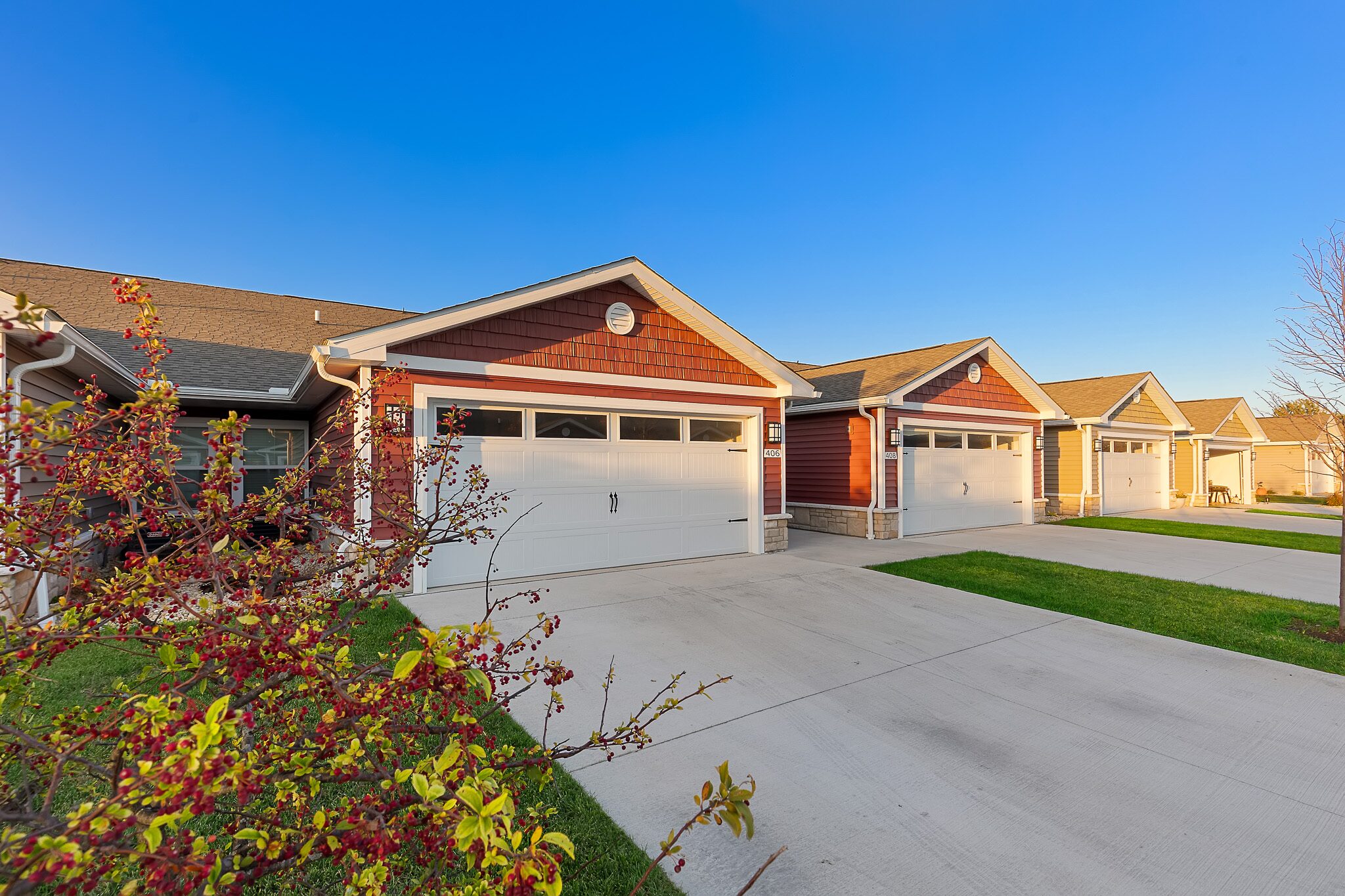Row of modern suburban homes with attached garages, neatly paved driveways, green lawns, and clear blue sky in a residential neighborhood.