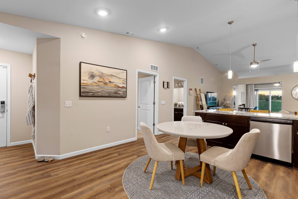 Open-concept dining and kitchen area with a round table and four chairs, dark cabinets, stainless steel appliances, and wood-style flooring. Artwork and bright lighting are visible.
