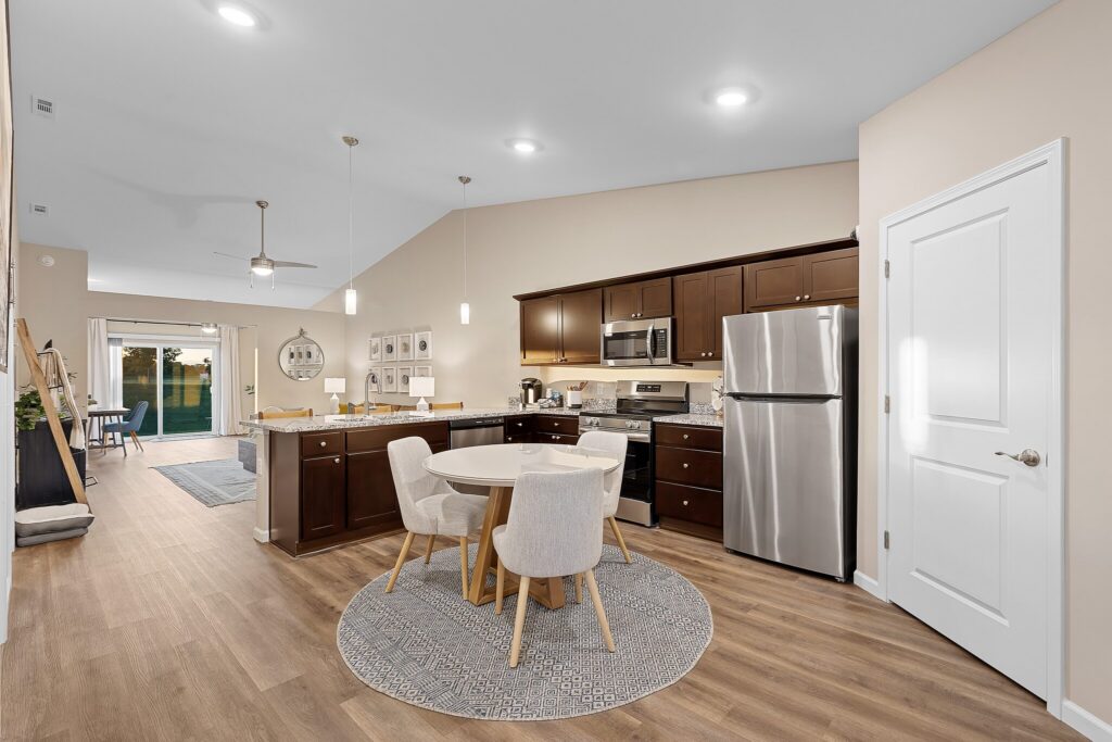 Modern open-concept kitchen and dining area with dark wood cabinets, stainless steel appliances, a round dining table, and light wood flooring. Living room visible in the background.