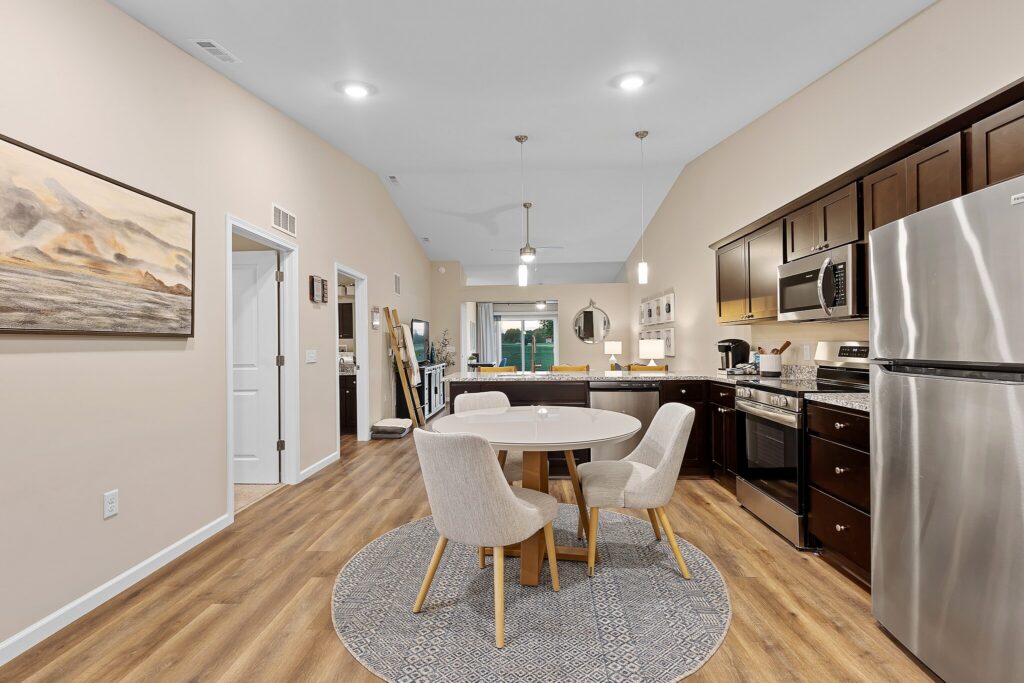 Modern open-concept kitchen and dining area with dark wood cabinets, stainless steel appliances, round dining table, and four beige chairs on a circular rug.