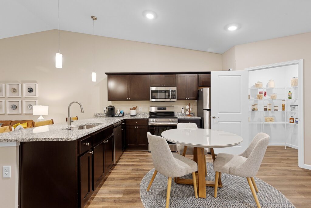 Modern kitchen with dark wood cabinets, stainless steel appliances, a round dining table with four chairs, and an open pantry with shelves stocked with food items.