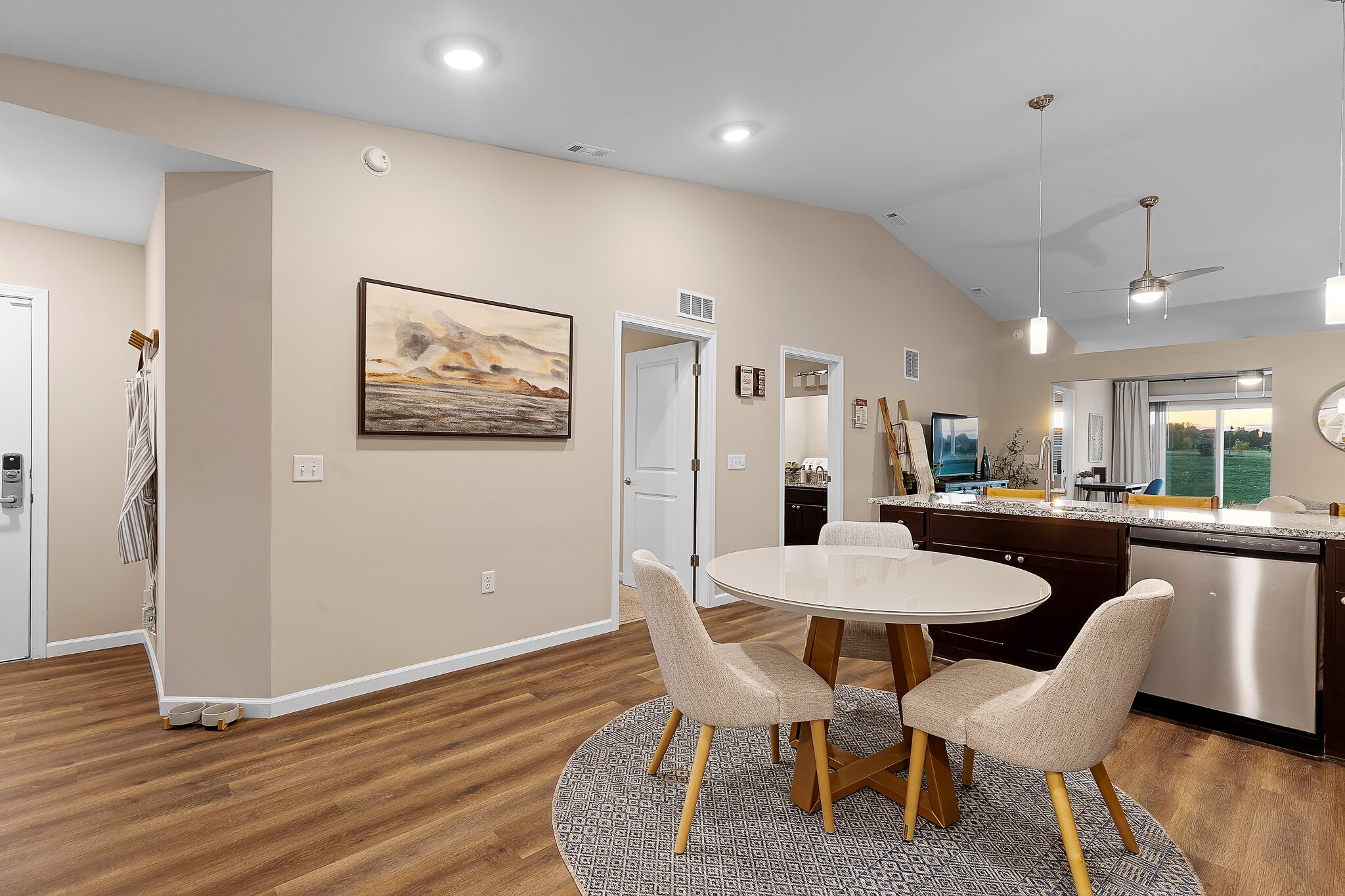 Open-concept dining and kitchen area with a round table and four chairs, dark cabinets, stainless steel appliances, and wood-style flooring. Artwork and bright lighting are visible.