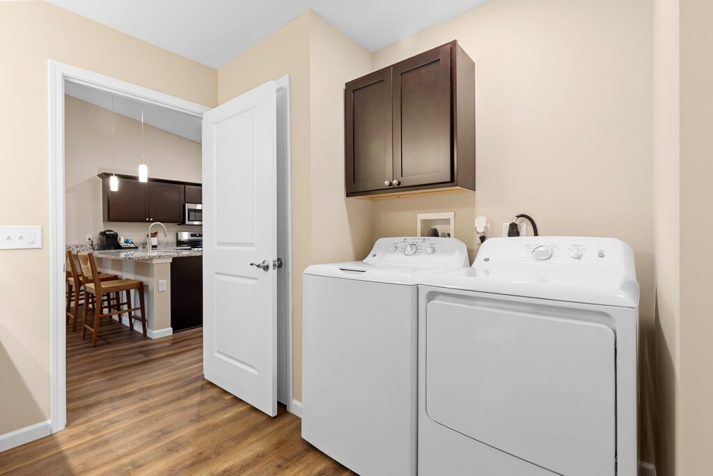 Laundry room with a white washing machine and dryer, brown cabinets above, and an open door leading to a kitchen and dining area with wood flooring.