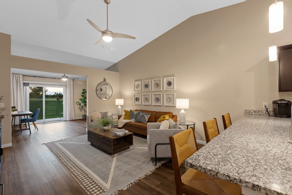 Open-concept living room with tan walls, brown sofa, armchair, coffee table, area rug, and dining area in the background near sliding glass doors to the outside.