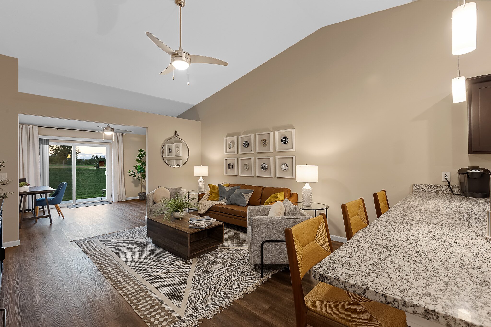 Open-concept living room with tan walls, brown sofa, armchair, coffee table, area rug, and dining area in the background near sliding glass doors to the outside.