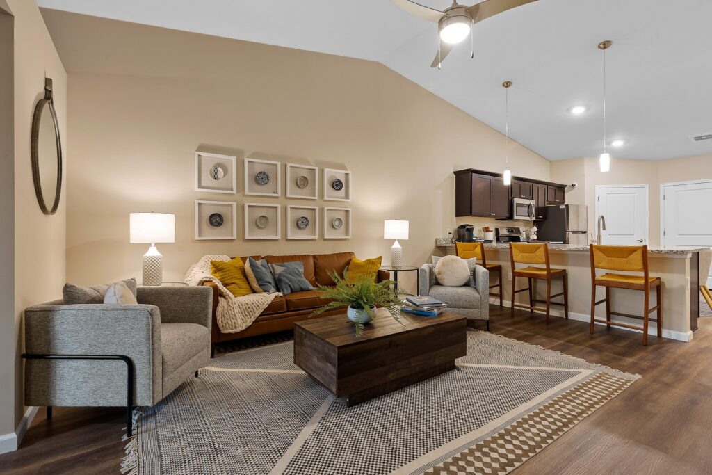 Modern open-concept living room and kitchen with neutral tones, a brown sofa, armchair, wooden coffee table, bar stools, and wall art above the couch.