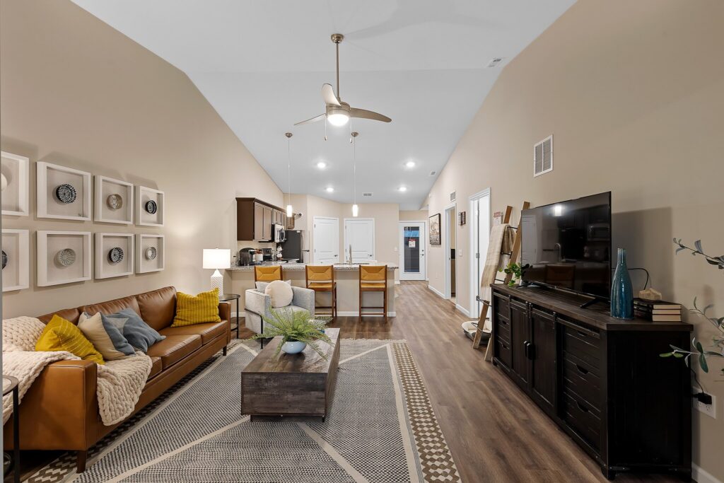 Open-concept living room and kitchen with brown leather sofa, wall art, TV on a black console, and a kitchen island with three stools under pendant lights.