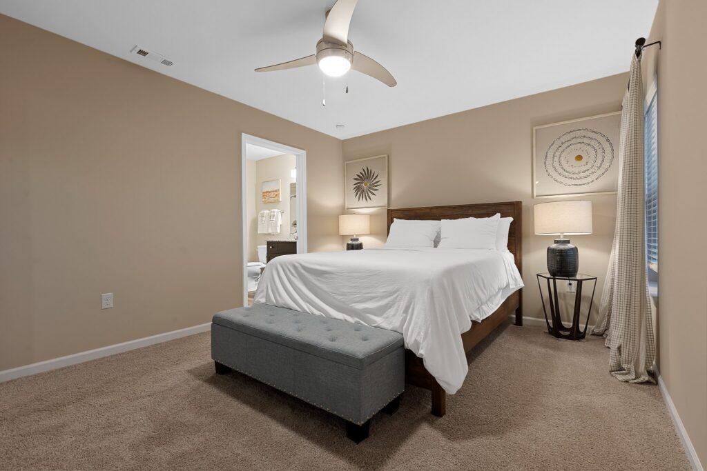 A neatly made bed with white bedding, a gray upholstered bench at the foot, two lamps on side tables, and beige walls in a carpeted bedroom with an open doorway to a bathroom.