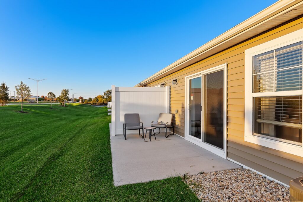 A small patio with two chairs and a table sits outside a tan house with sliding glass doors, next to a grassy lawn under a clear sky.