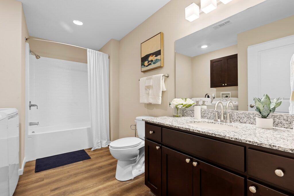 Modern bathroom with dark wood cabinets, granite countertop, white bathtub with shower curtain, toilet, large mirror, and decorative plants.