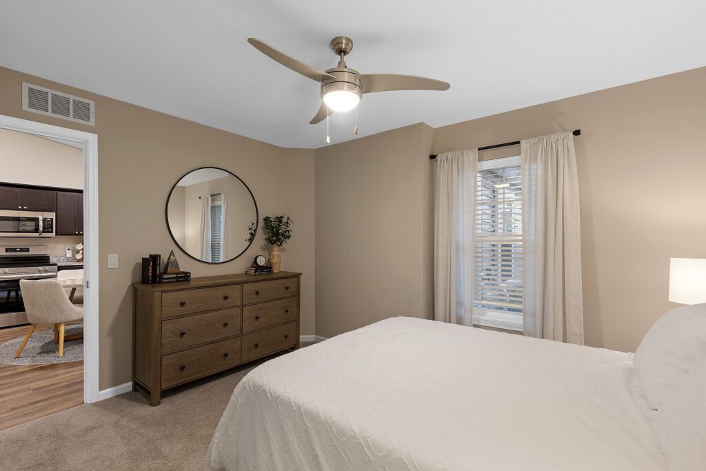 A bedroom with beige walls, a bed with a white comforter, a wooden dresser with a round mirror above it, and a window with white curtains. Ceiling fan with light is on.