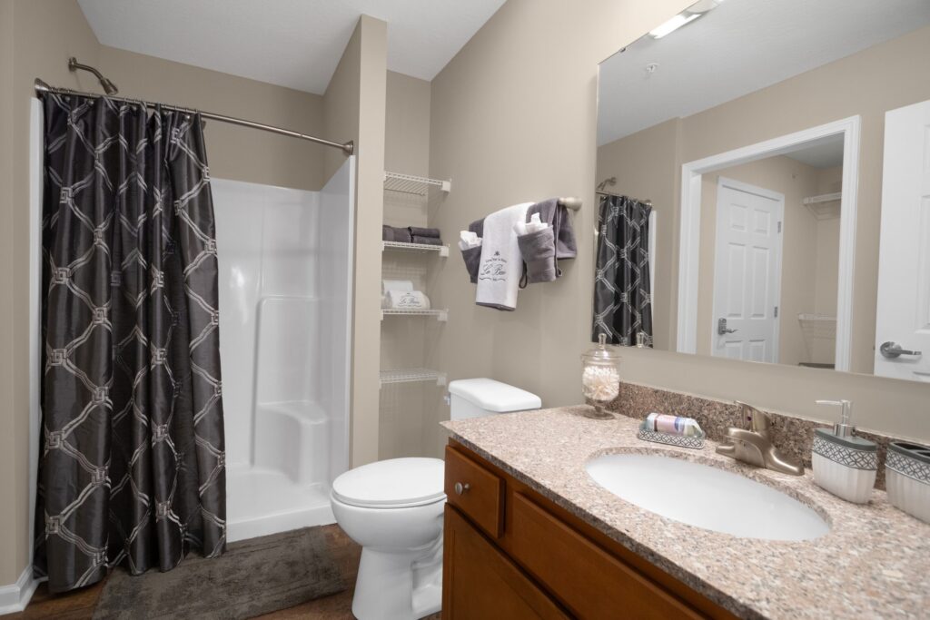 Bathroom with beige walls, a granite countertop vanity, toilet, shower with dark curtain, open shelving with towels, and a large mirror above the sink.