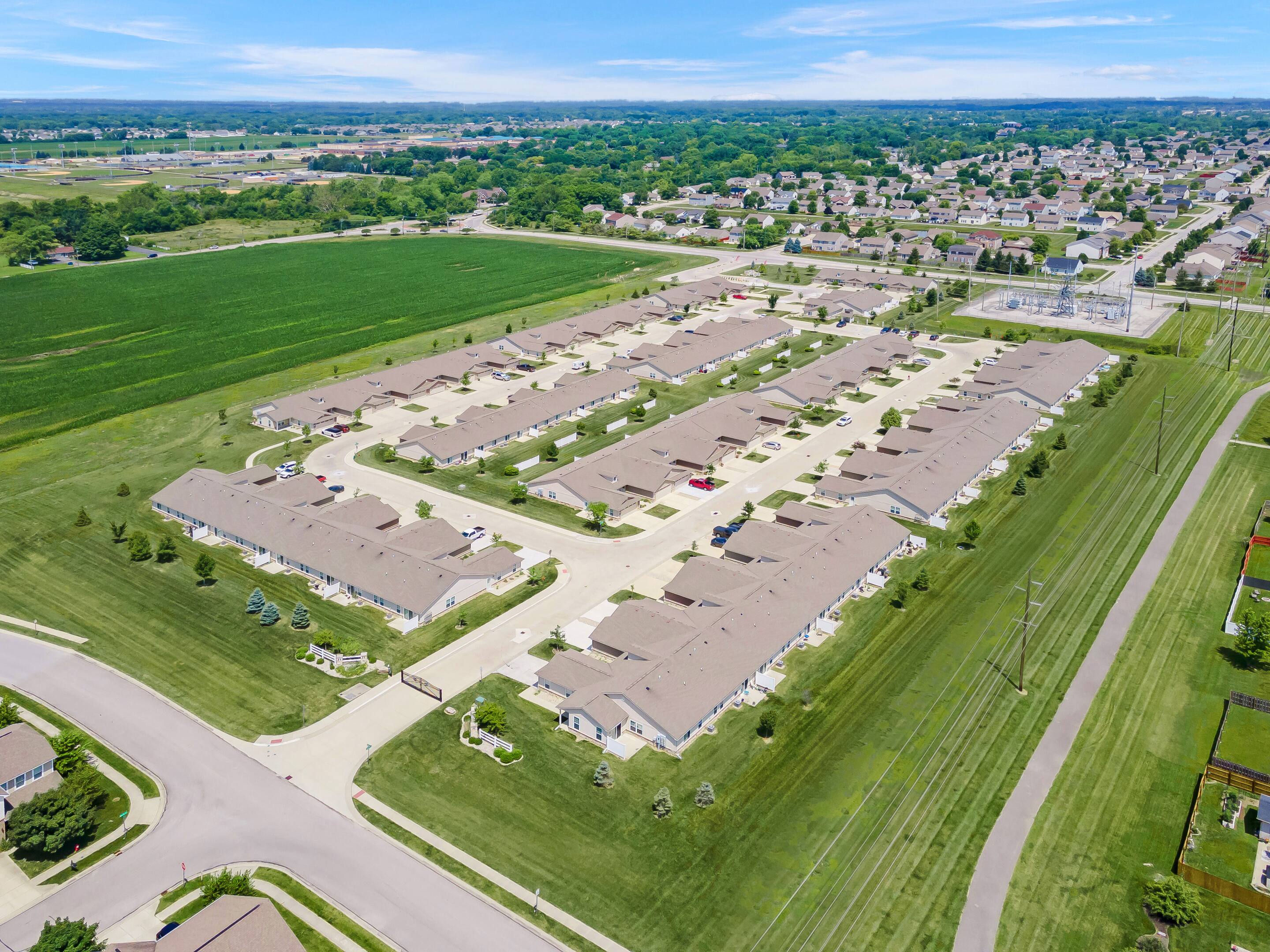Aerial view of a suburban residential neighborhood with rows of single-story houses, green lawns, nearby fields, and surrounding roads under a clear blue sky.