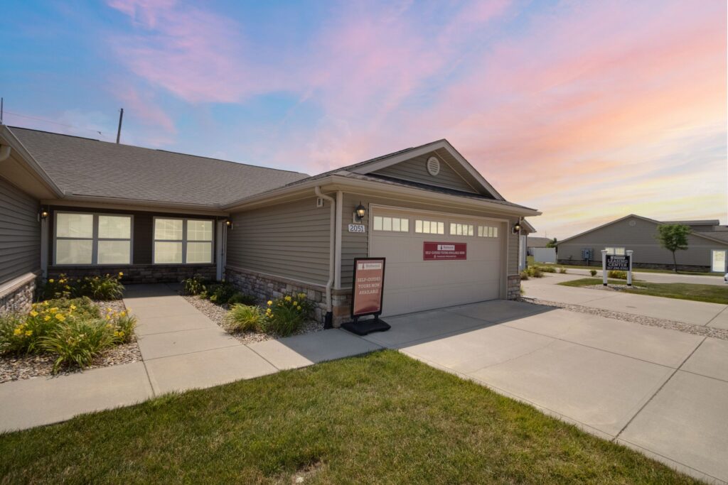 Single-story house with beige siding, a two-car garage, small front garden, and concrete walkway at sunset. Real estate signs are visible by the garage and across the street.