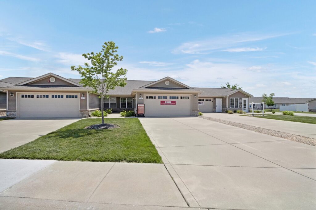 Single-story, attached residential homes with two-car garages, a small tree planted in the front yard, and wide concrete driveways on a clear day.