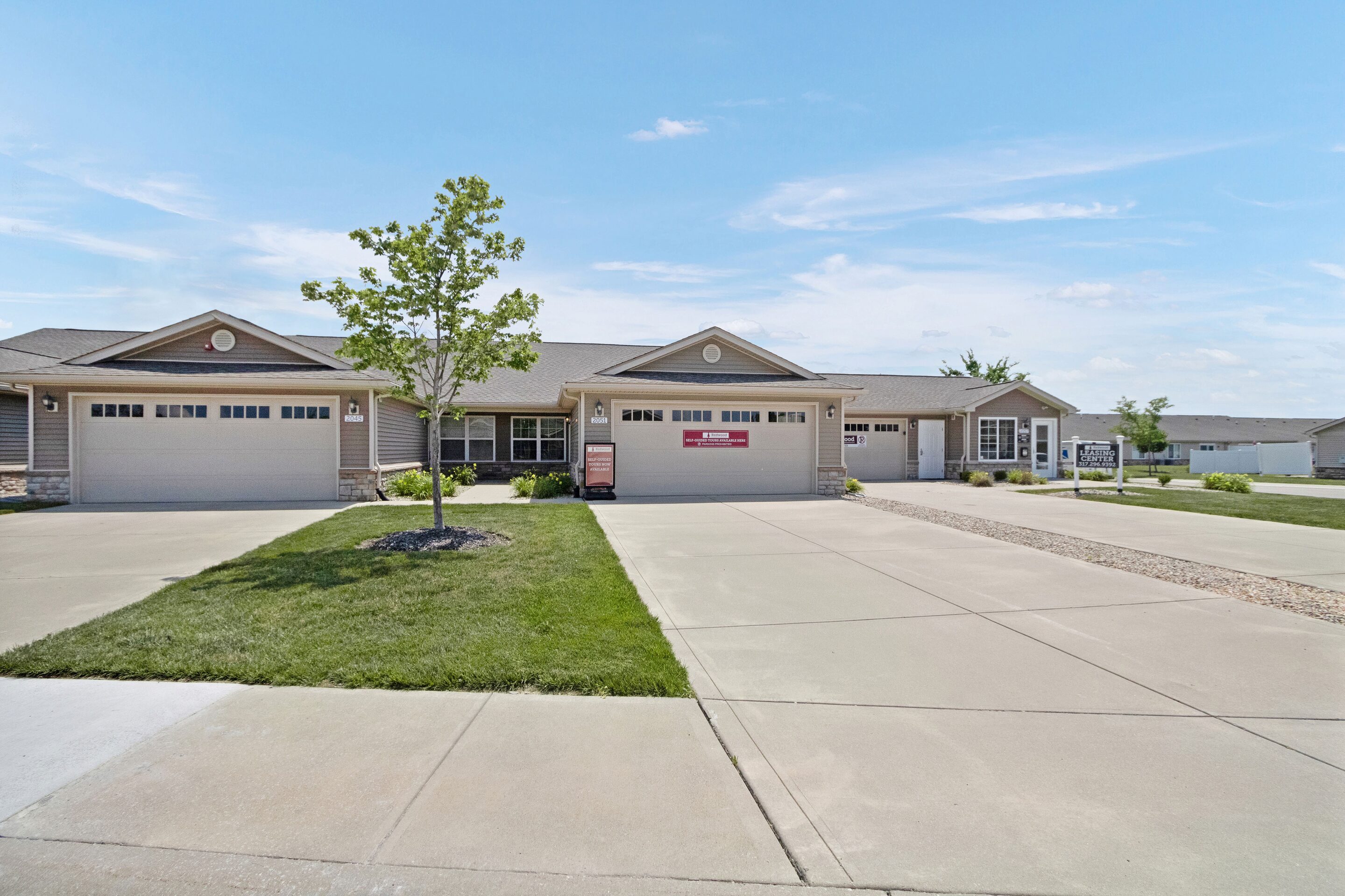 Single-story, attached residential homes with two-car garages, a small tree planted in the front yard, and wide concrete driveways on a clear day.