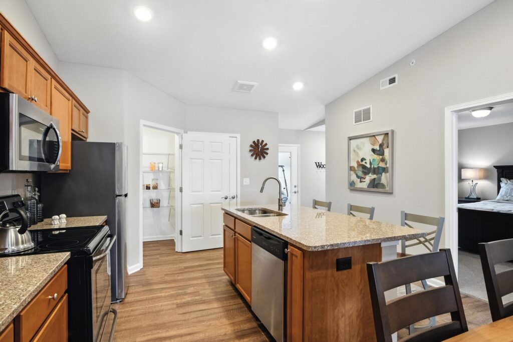 Modern kitchen with stainless steel appliances, granite island, wooden cabinets, and bar stools; pantry door open; bedroom visible through doorway.