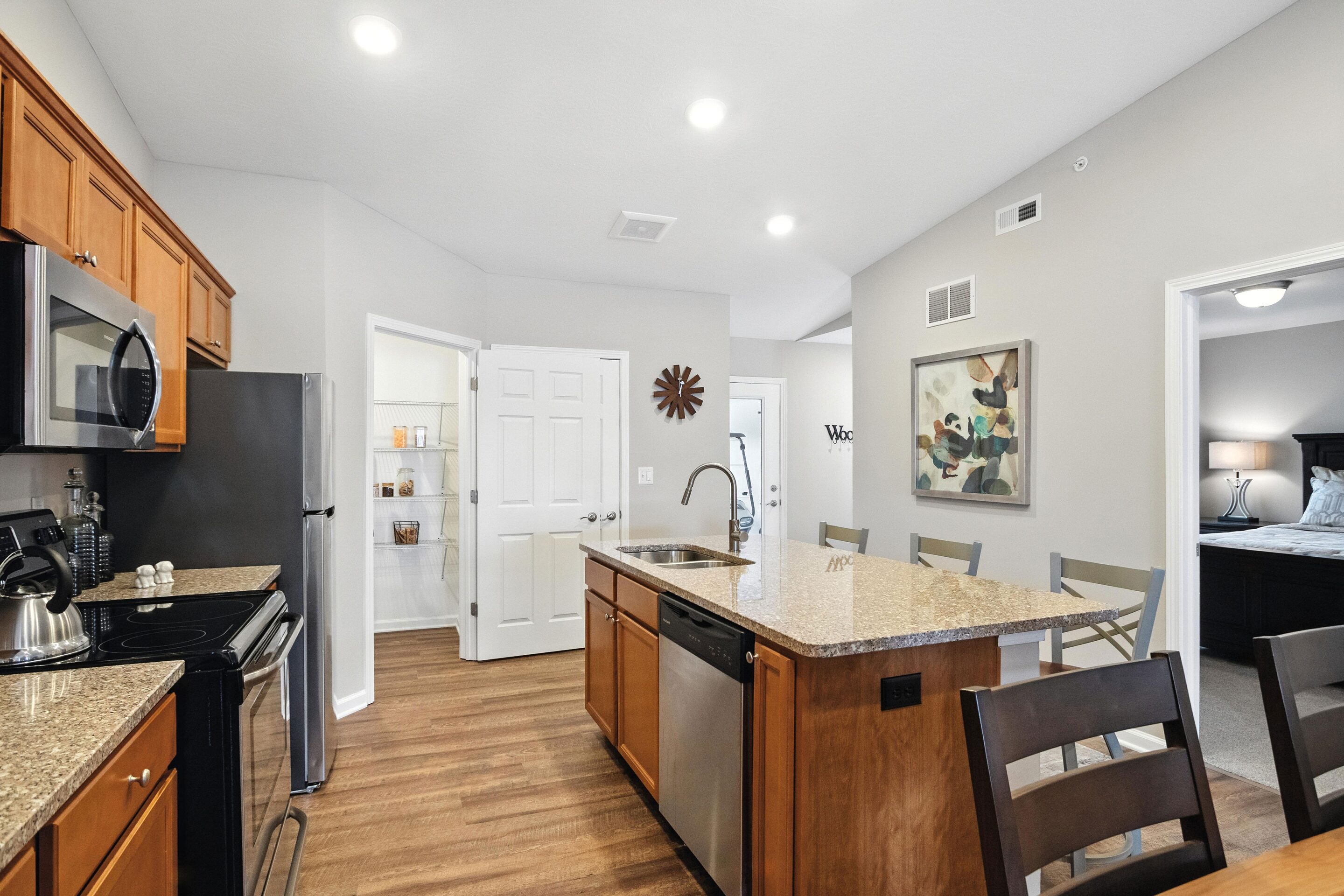 Modern kitchen with stainless steel appliances, granite island, wooden cabinets, and bar stools; pantry door open; bedroom visible through doorway.