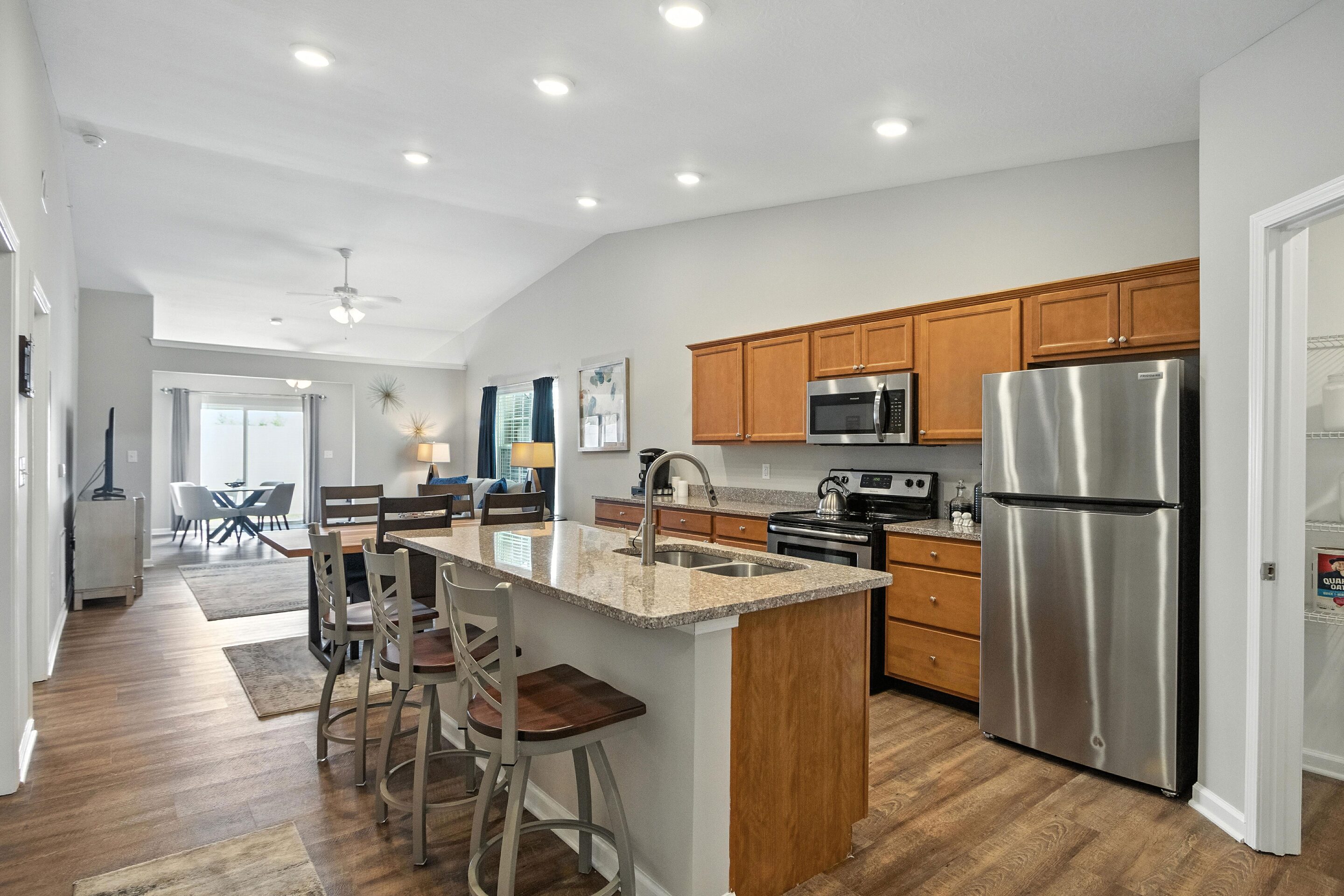 Modern kitchen with stainless steel appliances, wooden cabinets, an island with seating, and an open layout leading to a dining and living area.