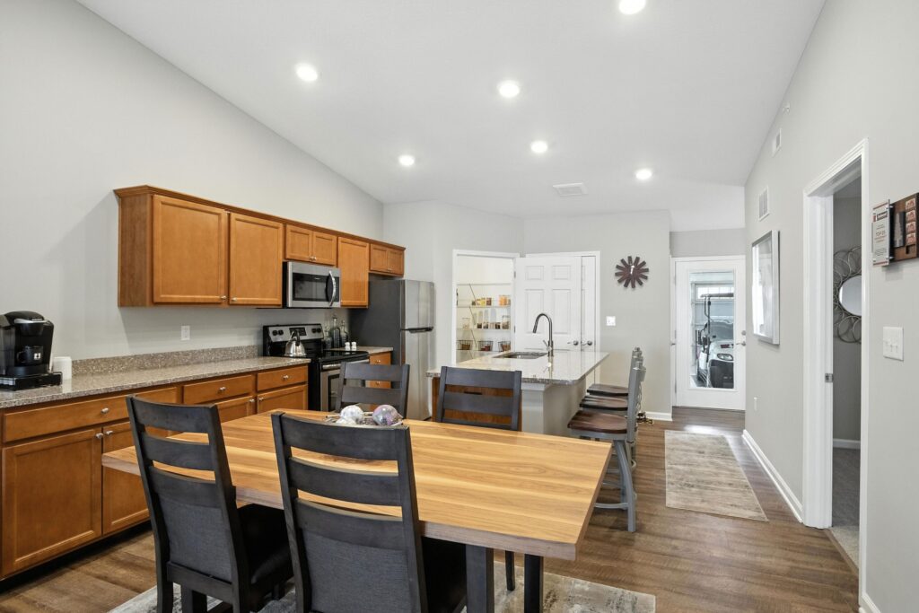 Modern kitchen and dining area with wooden cabinets, stainless steel appliances, a center island with a sink, and a wooden table with four chairs under recessed lighting.