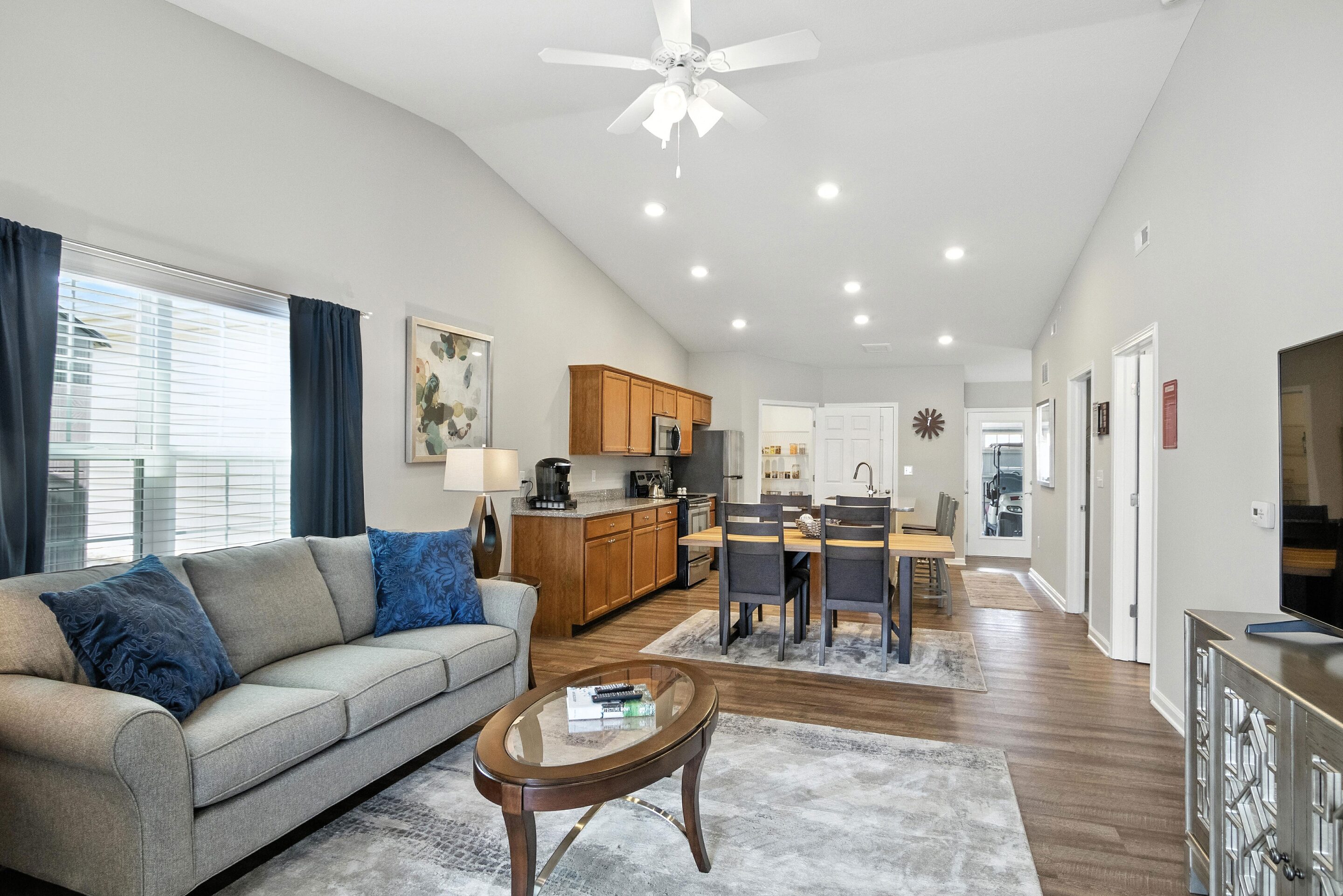 Open-concept living room and kitchen with gray sofa, wooden dining table, ceiling fan, and recessed lighting. Large windows and neutral decor create a bright, spacious atmosphere.