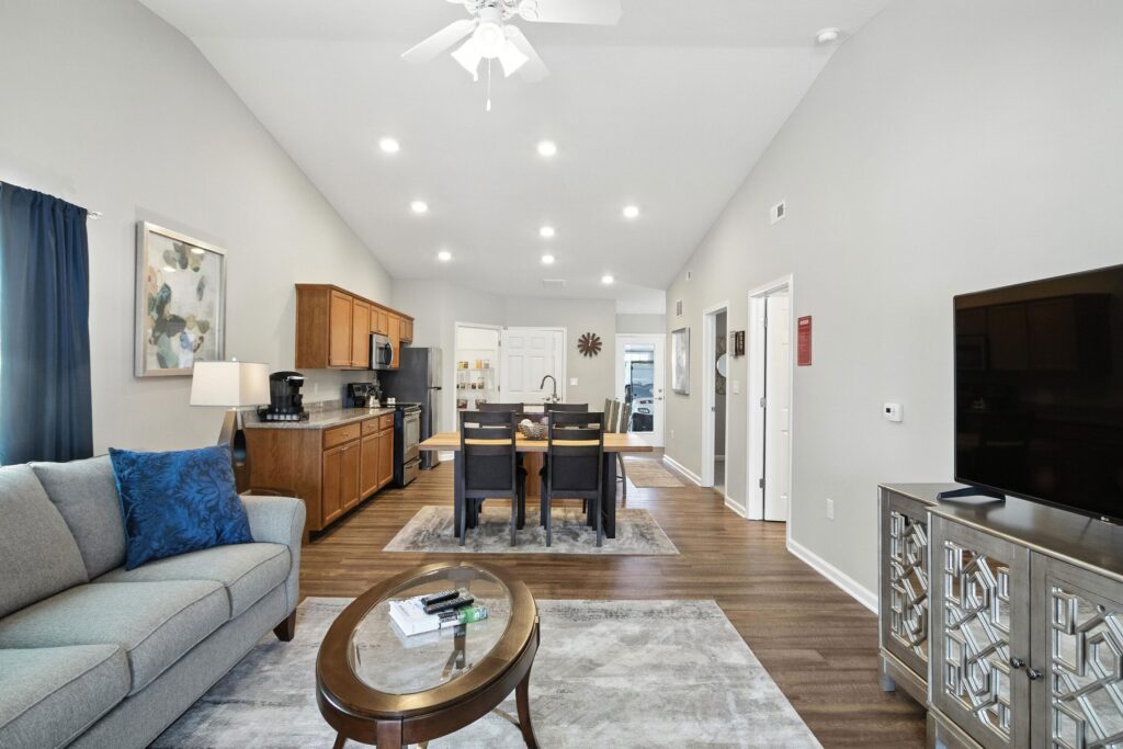 Open-concept living room and kitchen with gray sofa, wooden dining table, modern cabinetry, and a TV on a mirrored console. Ceiling fan and recessed lights are on.