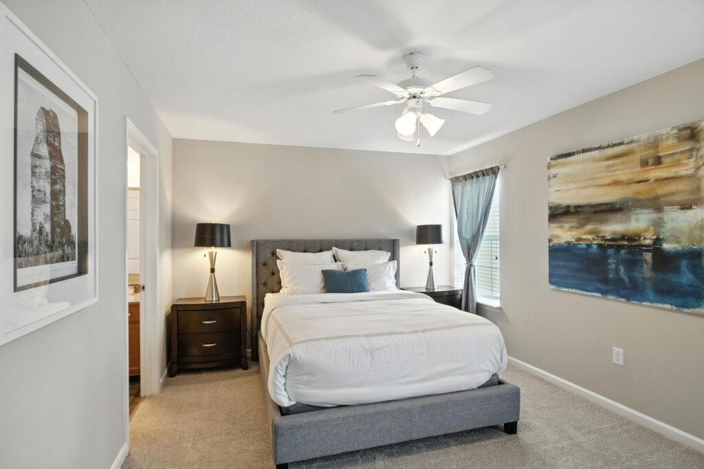 A neatly made bed with white bedding is centered between two dark nightstands with lamps in a neutral-toned bedroom with a ceiling fan and large wall art.