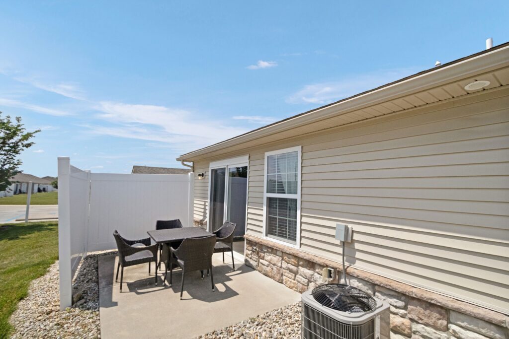 A small outdoor patio with a table and five chairs sits next to a beige house with siding and stone trim, adjacent to a white privacy fence and an air conditioning unit.