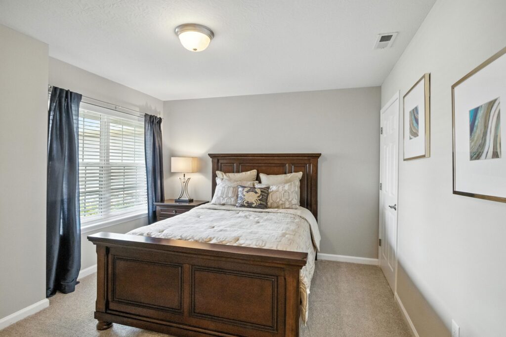 A neatly made bed with wooden frame in a simple bedroom, featuring a window with dark curtains, a nightstand with a lamp, and framed artwork on light-colored walls.