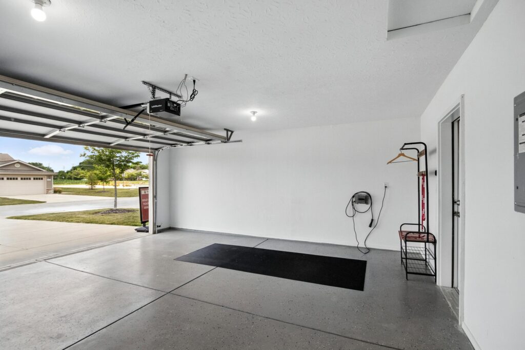 Empty two-car garage with white walls, concrete floor, open door, ceiling light, wall-mounted charger, black mat, and coat rack. Suburban houses visible outside.