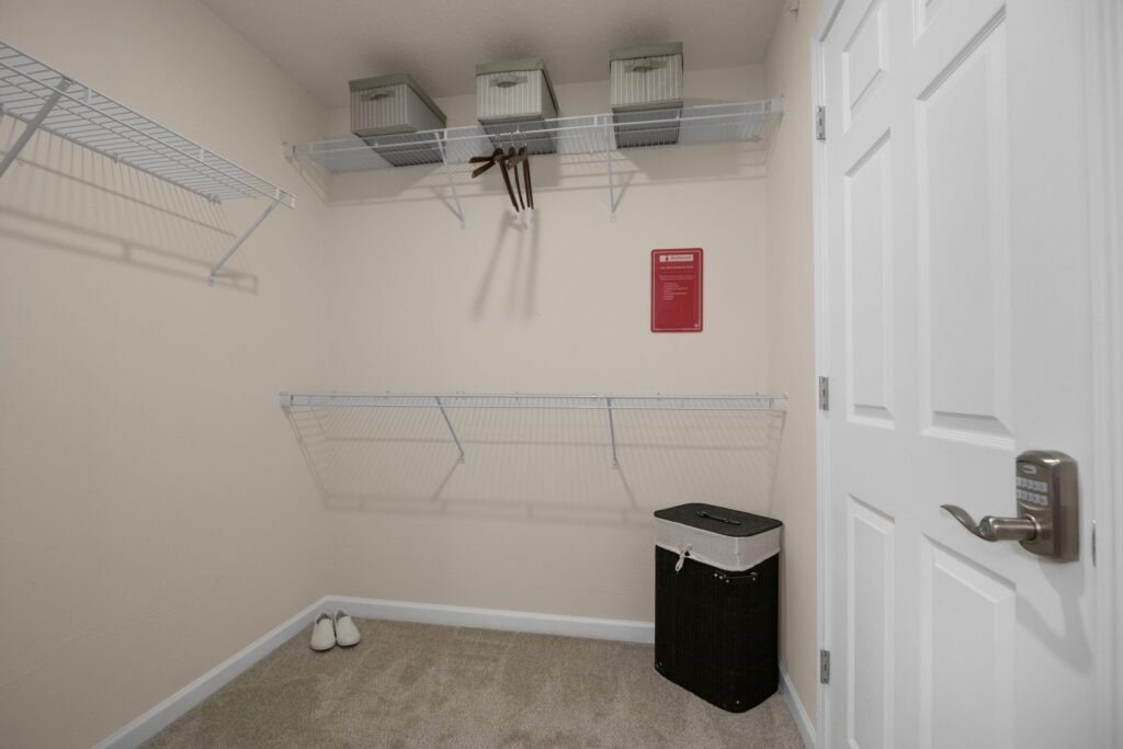 An empty walk-in closet with wire shelves, a few storage boxes, some hangers, a laundry hamper, and a pair of white shoes on the carpeted floor.
