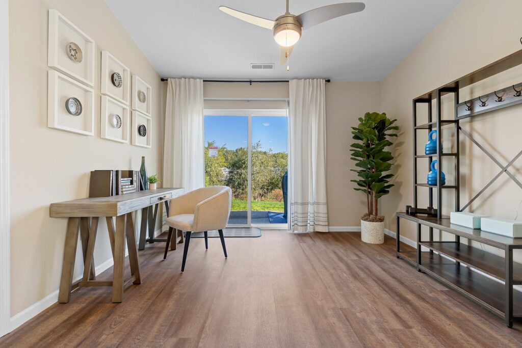 Modern home office with a wooden desk, beige chair, large plant, wall art, shelving unit, and sliding glass doors leading to a backyard with greenery.