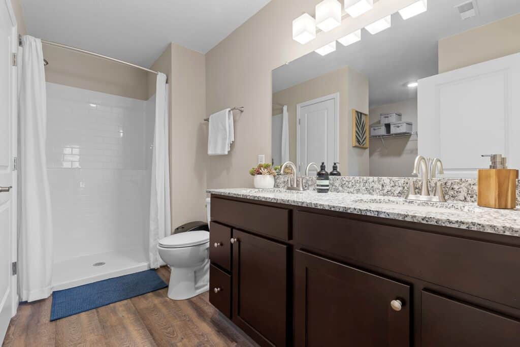 Modern bathroom with a granite countertop, double sink vanity, large mirror, walk-in shower with white curtain, towel rack, and wood-look flooring.