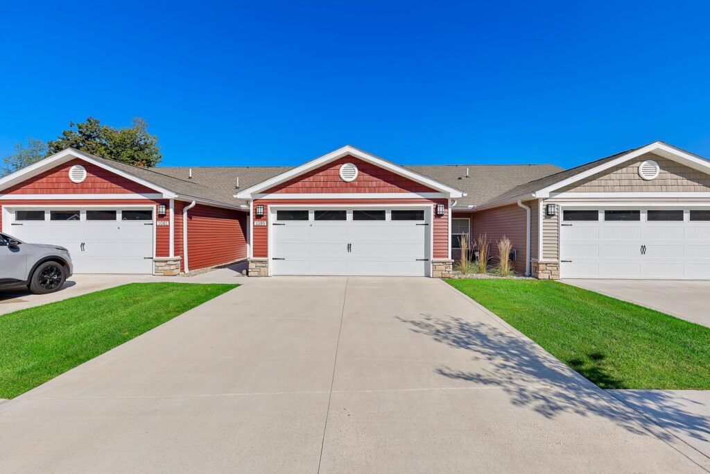 Three adjacent single-story townhouses with attached double garages, concrete driveways, red and beige siding, and a blue sky overhead.