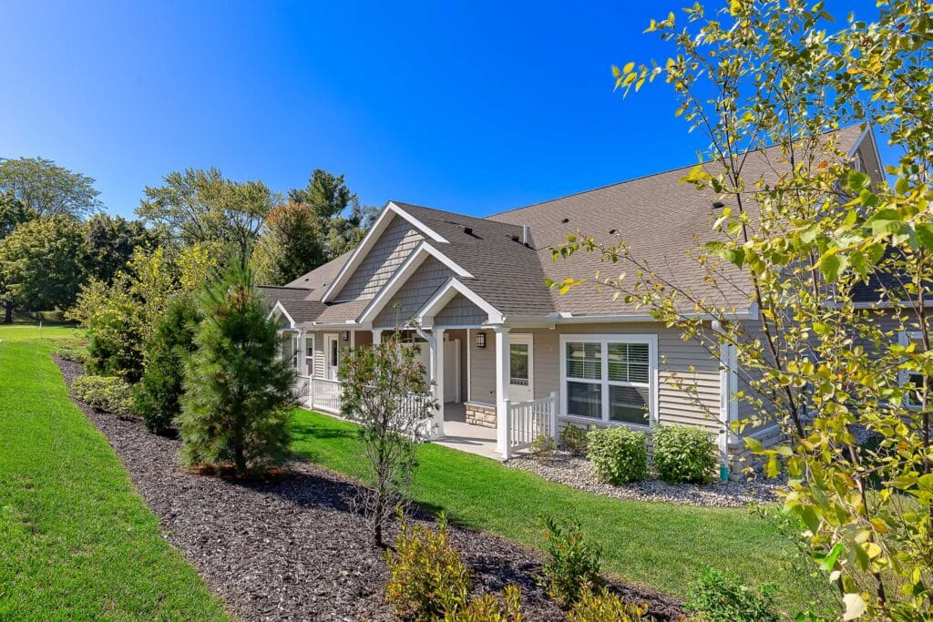 Single-story house with beige siding, white trim, and gabled roof, surrounded by a well-maintained lawn, shrubs, and trees under a clear blue sky.