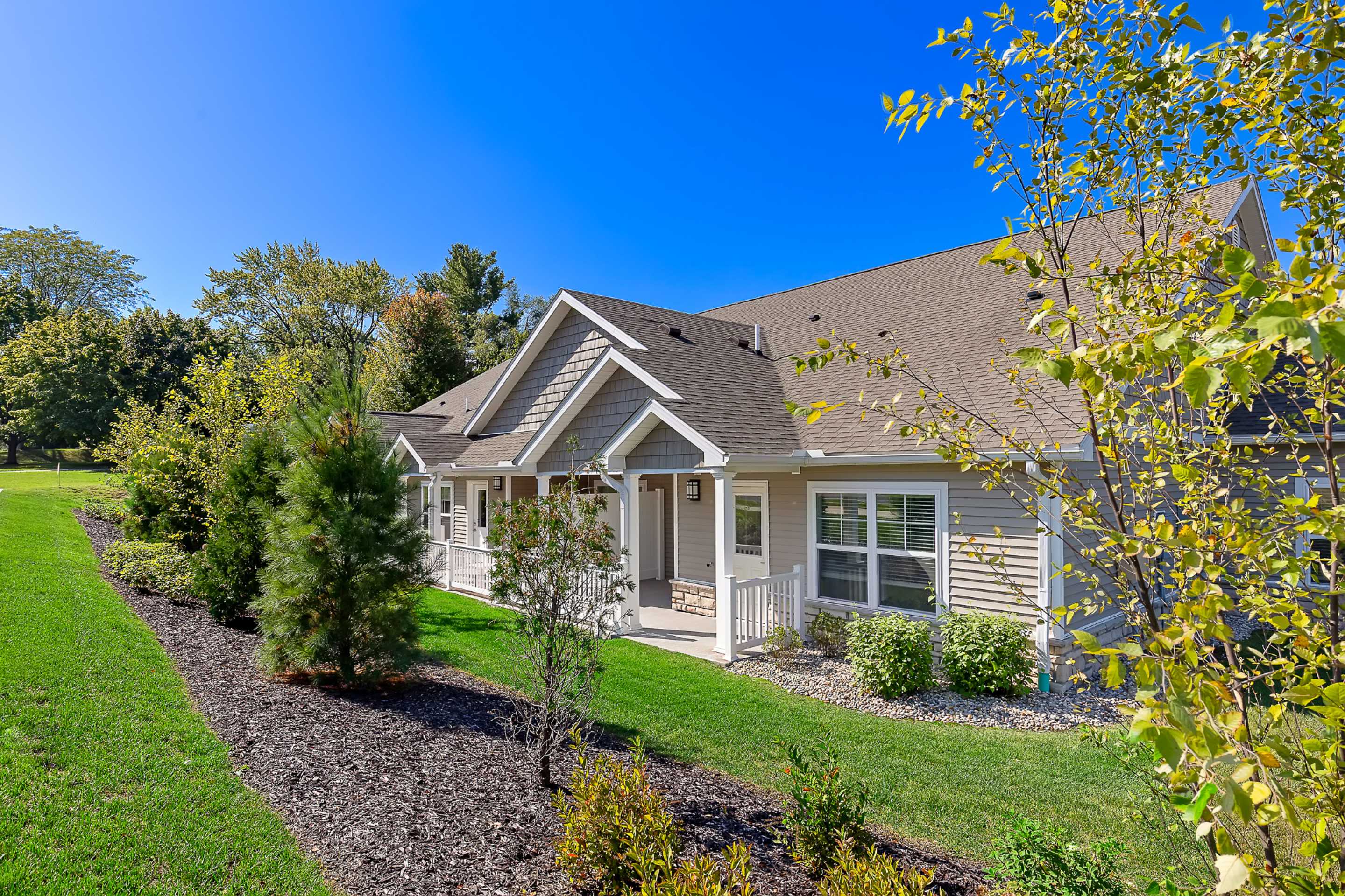 Single-story house with beige siding, white trim, and gabled roof, surrounded by a well-maintained lawn, shrubs, and trees under a clear blue sky.